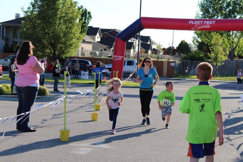A young girl runs alongside an adult pacer and another young runner wearing a race bib, as they near a course arch during the SoleKIDs 5K