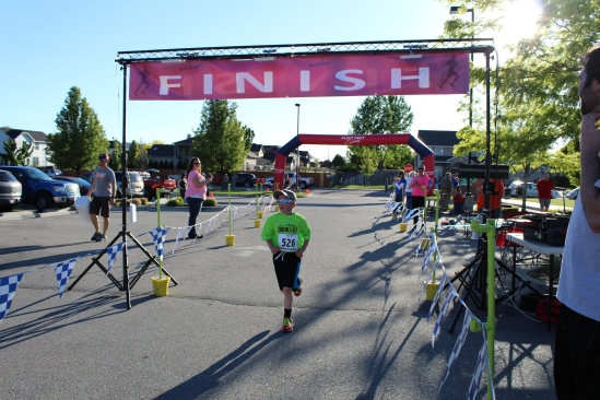A young runner in a lime green race shirt crosses under a red FINISH banner at the SoleKIDs 5K