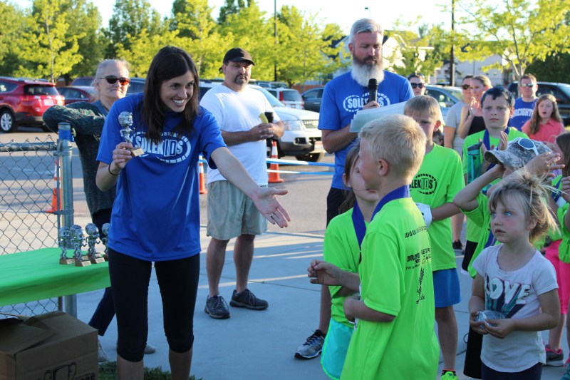 A SoleKIDs coach hands a trophy to a young runner in a lime green shirt as other kids wait in line at the awards table