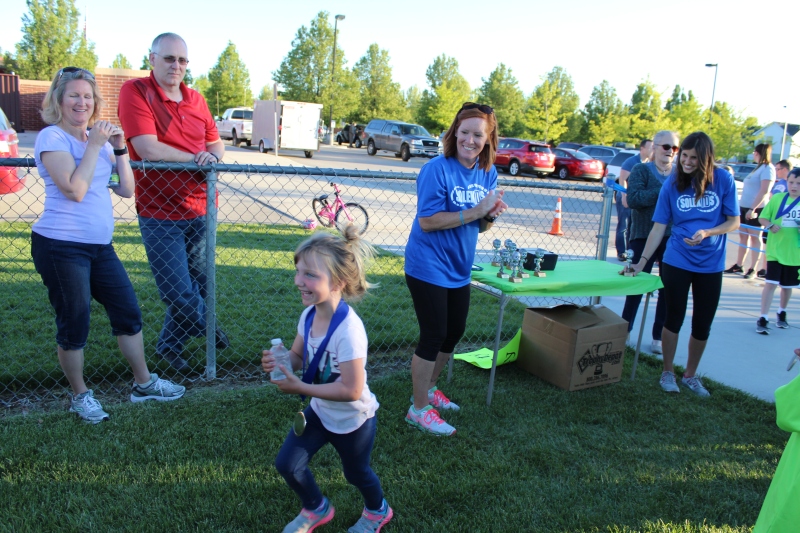 A young girl wearing a finisher's medal smiles and runs across the grass while adults clap and cheer around her