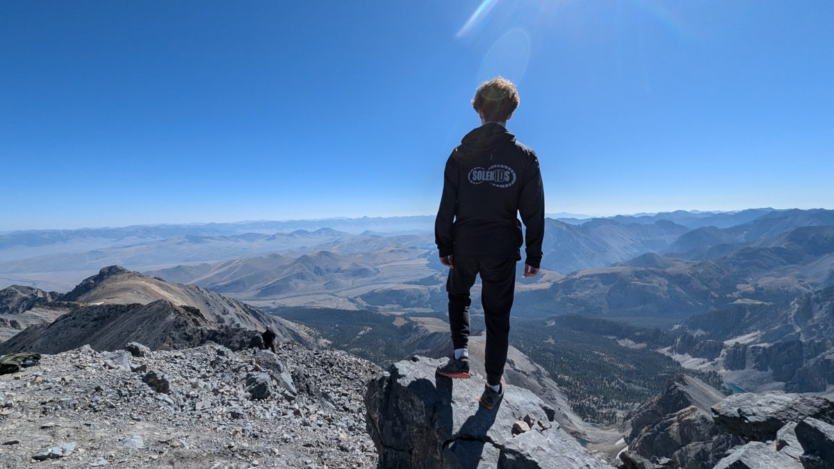A SoleKIDs runner in a SoleKIDs hoodie standing on a mountain summit, looking out over the Idaho backcountry.